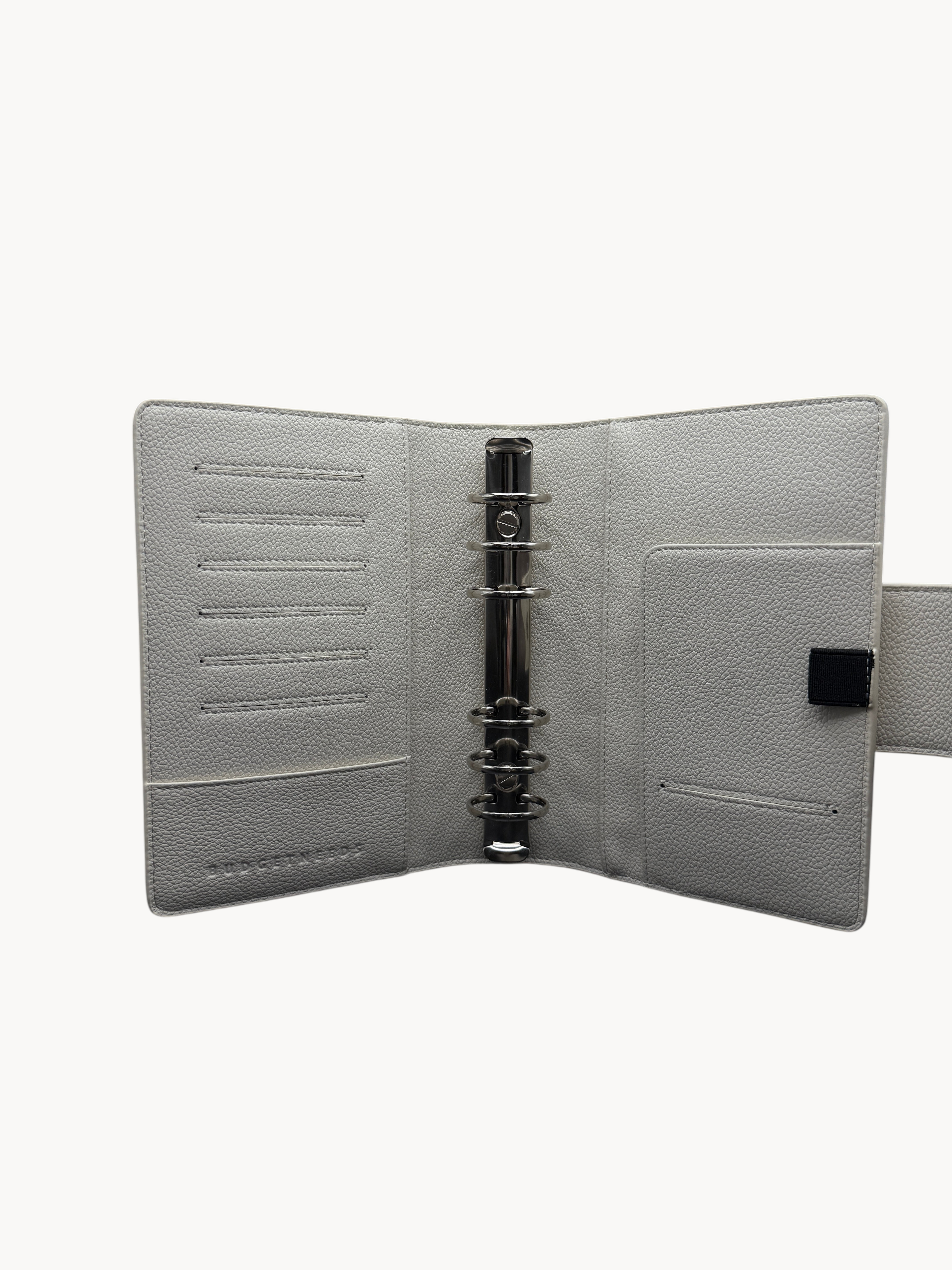BUDGETNEEDS ™ A6 MiniFlap ring binder in cream with silver hardware, pen holder, and card slots on a black background.